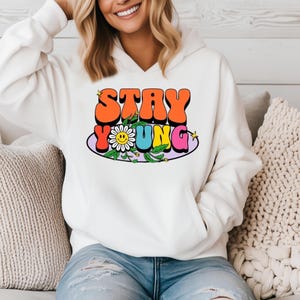 May include: A white hooded sweatshirt featuring a graphic print on the chest with the text STAY YOUNG in bold, colorful, bubble-style letters. The design includes a white daisy flower with a yellow smiley face center and small yellow stars.
