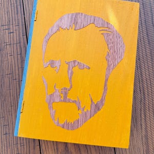 May include: A wooden notebook with a yellow cover featuring a cutout portrait of a man with a beard. The spine is blue, and the notebook is open. The background is a wooden surface.