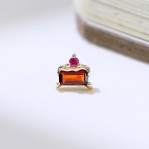 May include: A small, gold-colored jewelry piece with a rectangular, orange-red gemstone and a smaller, round, red gemstone on top. The piece is set against a white background with a partially visible stack of paper.