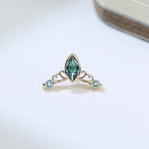 Könnte beinhalten: Ein zierliches, goldfarbenes Schmuckstück in Form einer Tiara. Der zentrale Edelstein ist ein oval geschliffener teal-farbener Stein, flankiert von zwei kleineren, runden, hellblauen Edelsteinen. Das Design weist filigrane Details auf.