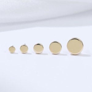 May include: A collection of six gold-colored, round stud earrings of varying sizes, arranged in a row. The earrings are displayed on a white surface, with a soft, white background.