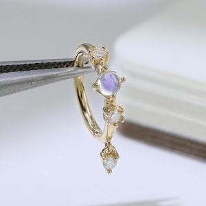 Può includere: Un orecchino a cerchio dorato con una pietra di luna e tre piccole gemme trasparenti. L'orecchino è tenuto da una pinzetta argentata su sfondo bianco. La pietra di luna ha una morbida lucentezza iridescente.