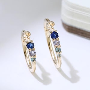 May include: A pair of gold hoop earrings, each embellished with a sequence of colourful gemstones. The earrings showcase a mix of blue, purple, and green stones, creating a vibrant design. The earrings are set against a white background.