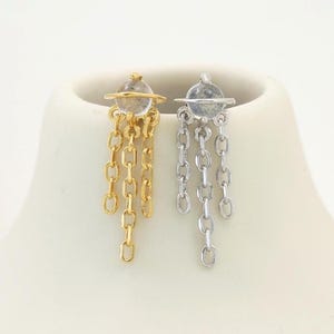 Puede incluir: Un par de pendientes, uno dorado y otro plateado, cada uno con una pequeña parte superior circular y eslabones de cadena colgantes. Los pendientes se muestran sobre un busto de maniquí blanco sobre un fondo blanco.