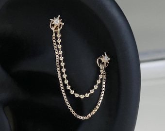May include: A gold-toned ear piercing jewelry piece featuring two star-shaped studs connected by two delicate, dangling chains. The upper stud is linked to the lower stud by a beaded chain and a box chain, creating a layered, draped effect.
