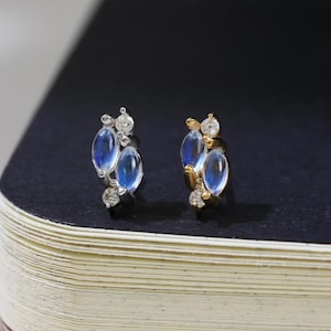 May include: A pair of delicate stud earrings featuring three oval, light blue gemstones and small clear accent stones. The earrings are set in a gold or silver metal. The earrings are displayed on a dark blue surface.