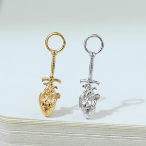 May include: Two heart-shaped pendant earrings, one gold-toned and one silver-toned, each with a sword-like design. The earrings are displayed on a white surface, showcasing their intricate details and contrasting colors.