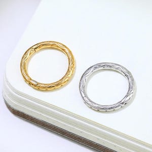 Może przedstawiać: Dwa teksturowane metalowe pierścienie, jeden złoty i jeden srebrny, są wyświetlane na białej powierzchni. Pierścienie mają okrągły kształt ze szczegółowym, wzorzystym wzorem. Złoty pierścień jest po lewej stronie, a srebrny po prawej.