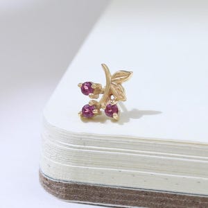 May include: A gold-colored stud earring featuring a leaf design with three small, round, red gemstones. The earring is placed on a white surface, with a soft shadow to the right.
