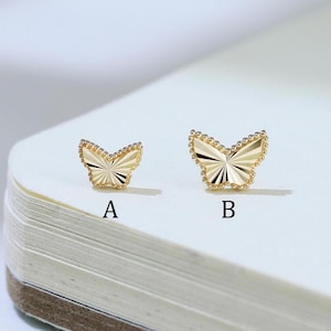 May include: Two gold-colored butterfly-shaped stud earrings, labeled A and B, featuring a textured, sunburst-patterned surface with a beaded edge. The smaller earring is marked with the letter A, and the larger earring is marked with the letter B.