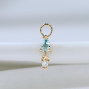 May include: A gold charm featuring a small, pear-shaped light blue gemstone suspended above a tiny, round, clear white gemstone. The charm includes a circular jump ring at the top for attachment, showcasing a delicate and refined metal construction.