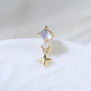 Könnte beinhalten: Zarter goldener Ohrring mit einem quadratischen, schillernden Edelstein, einem kleinen Diamanten und einem sternförmigen Anhänger. Der Ohrring ist vor einem weichen, weißen Hintergrund platziert, der sein elegantes Design hervorhebt. Ein funkelndes Schmuckstück.