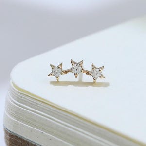 May include: A delicate gold-toned earring featuring three star-shaped designs, each adorned with small, sparkling gemstones. The earring is positioned on a stack of white paper, with soft shadows in the background. The stars are evenly spaced and add a touch of celestial charm.