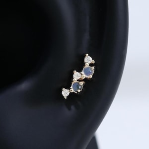 May include: A gold-toned ear stud featuring a curved, cluster design with two round, light blue stones and three small, clear white accent stones set in a delicate metal frame, positioned on a dark, smooth ear-shaped display form.