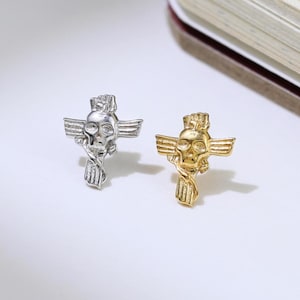 May include: Two skull-shaped stud earrings, one silver and one gold, each with wings and a twisted design below. The earrings are displayed on a white surface, next to a book.