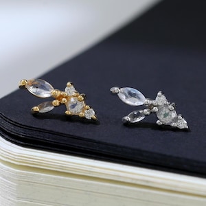 Puede incluir: Un par de delicados pendientes de botón con un diseño floral. Cada pendiente presenta un grupo de pequeñas piedras preciosas transparentes engastadas en metal dorado y plateado. Los pendientes se muestran sobre una superficie negra.