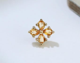 May include: A gold-colored stud earring with a four-petal flower design. Each petal features a light orange gemstone. The earring is set against a white background with a shadow.