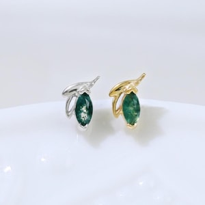 May include: Two stud earrings, one silver and one gold, each with a green gemstone in an olive leaf design. The earrings are presented on a white surface, highlighting their delicate craftsmanship and elegant style.