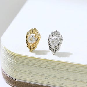 May include: Two feather-shaped stud earrings, one gold-toned and one silver-toned, each featuring a central clear gemstone. The earrings are displayed on a white surface, showcasing their intricate design and sparkling details. Jewelry.