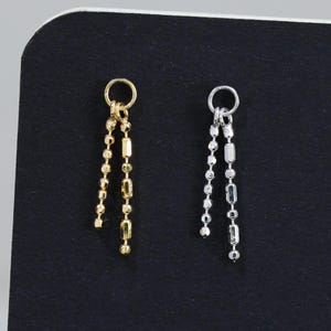 May include: Two dangle earrings, one gold-toned and one silver-toned, are displayed against a dark background. Each earring features a small circular top with multiple chain strands of varying lengths, adorned with small rectangular and spherical beads.