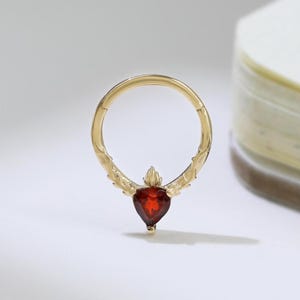Peut inclure: Une bague dorée ornée d'une pierre précieuse rouge foncé en forme de cœur. La bague présente des détails décoratifs sur les côtés et une fermeture à charnière. La pierre précieuse mesure environ 0,6 cm de long. La bague est posée sur un fond blanc.