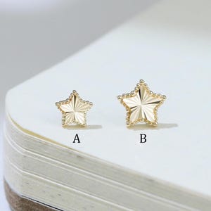 May include: Two gold-colored star-shaped earrings, labeled A and B, are displayed on a white surface. The earrings have a textured design with a starburst pattern. The earrings are likely for pierced ears.