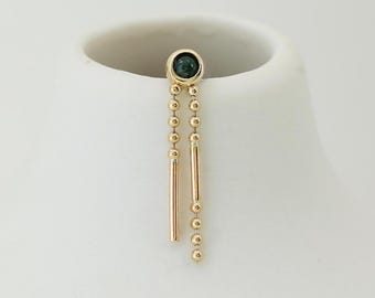 May include: A gold-colored earring with a dark green gemstone at the top. Two gold chains hang down, one with small gold beads and the other with a straight gold bar. The earring is displayed on a white surface.