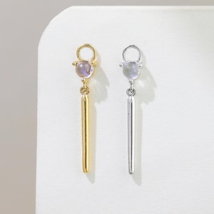 Puede incluir: Un par de pendientes, uno dorado y otro plateado, cada uno con una pequeña gema iridiscente en la parte superior y un colgante de barra vertical. Los pendientes se muestran sobre un fondo blanco.