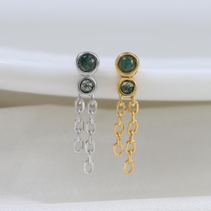 Puede incluir: Dos pares de pendientes colgantes, uno plateado y otro dorado. Cada pendiente tiene dos tachuelas de piedra verde y cadenas colgantes. Se presentan sobre una superficie blanca, destacando su diseño y contraste.
