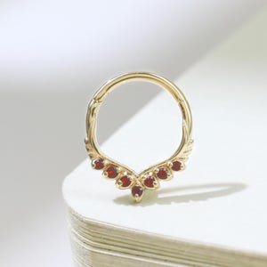 May include: A gold-colored septum ring with a decorative, downward-pointing design. The design is adorned with small, round, red gemstones. The ring is set against a white background, with soft shadows.