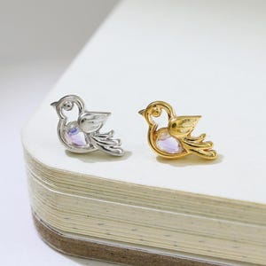 以下が含まれることがあります： 鳥の形をしたスタッドピアス2つ。1つはシルバー色、もう1つは金色です。どちらも真珠光沢のある中心部があり、白い表面に置かれています。ピアスは小さく繊細で、普段使いに適しています。