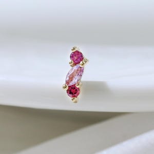 Puede incluir: Un pendiente dorado con tres piedras preciosas rosas. Las piedras superior e inferior son redondas, mientras que la piedra central es ovalada. El pendiente está sobre un fondo blanco.