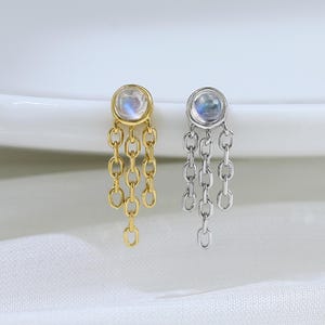 Op de afbeelding: Twee oorknopjes, één goudkleurig en één zilverkleurig, elk voorzien van een ronde maansteen cabochon in een zetting. Drie fijne, korte kettinkjes hangen aan de onderkant van elke ronde zetting, wat zorgt voor een gelaagd, hangend effect.