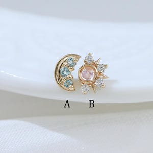 以下が含まれることがあります： AとBのラベルが付いた2つのゴールドスタッドピアス。ピアスAは3つの水色の石がセットされた三日月型。ピアスBは中央の丸い石を小さな透明な石が囲む太陽のデザイン。どちらもゴールドトーンの金属製です。
