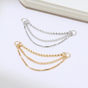 May include: Two sets of delicate chain earrings. One set is gold-toned, the other is silver-toned. Each earring features three strands of varying chain styles, including beaded and flat link chains. The earrings are displayed on a white surface.