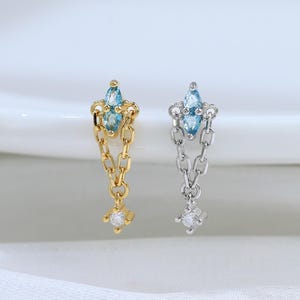 May include: Two dangle earrings, one gold-toned and one silver-toned, each featuring a marquise-cut light blue gemstone at the top, a short delicate chain link section, and a small round clear gemstone at the bottom of the dangling chain.