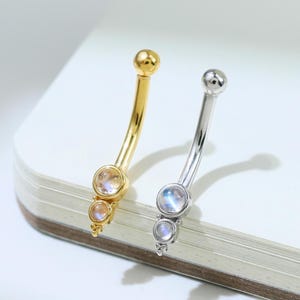 May include: Two curved belly button rings, one gold and one silver, each with a small, round gemstone at the top and two smaller gemstones at the bottom. The gemstones have a pearlescent, iridescent quality.