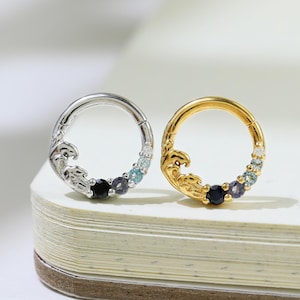 May include: Two circular earrings, one silver and one gold, each featuring a wave design and small, colorful gemstones. The silver earring has a black gem, while the gold earring has a dark blue gem. Both earrings have light blue and purple gems.