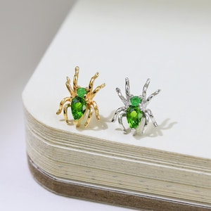 Puede incluir: Dos pendientes con forma de araña. Uno es dorado con una gema verde, el otro es plateado con una gema verde. Un broche de araña dorado está en el fondo. Los pendientes están sobre una superficie blanca.