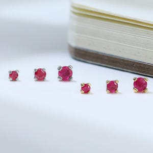 May include: A collection of small, round ruby earrings in varying sizes. The earrings are set in silver and gold settings, displayed on a white surface next to a partially visible notebook. The rubies are a vibrant red color.