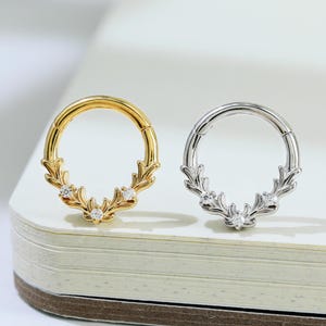 May include: Two hoop earrings, one gold-toned and one silver-toned, each featuring a decorative leaf design with small, sparkling gemstones. The earrings are displayed on a white surface, showcasing their intricate details and elegant style.