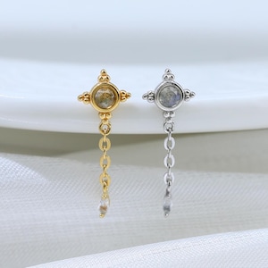 May include: Two earrings, one gold-toned and one silver-toned, each featuring a four-pointed star design with a central gemstone. A delicate chain hangs from each star. The earrings are displayed on a white surface.