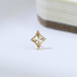 May include: A small, gold stud earring with a starburst design. The earring has a four-pointed star shape with textured detailing and a beaded border. The earring is set against a white background with a notebook in the background.