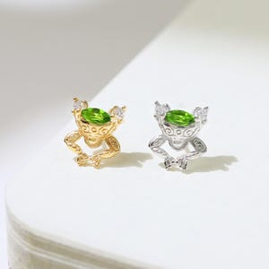 May include: Two small, detailed frog-shaped earrings. One is gold-toned, the other silver-toned. Each frog has a large, round, green gemstone set in its head. The earrings are displayed on a white surface.