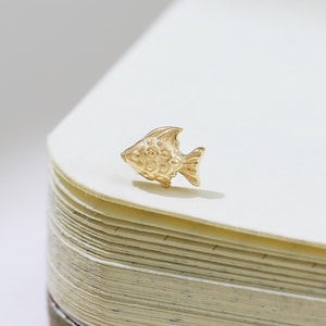 Pode incluir: Um pequeno pingente em forma de peixe dourado. O pingente está sobre o canto de um caderno aberto com páginas creme. O peixe tem escamas e barbatanas detalhadas.