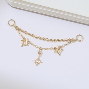May include: A delicate gold-colored jewelry piece featuring a chain with small beads and three dangling star-shaped charms. A smaller star charm with a central gem hangs below the others. The piece has two small loops on each end.