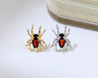 May include: Two spider-shaped earrings. One is gold-toned with a red center stone, the other is silver-toned with a red center stone. The earrings have detailed legs and a small, round body. The background is white.