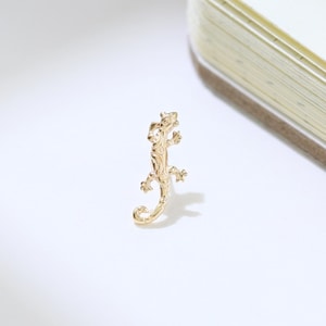 May include: A small, gold-colored lizard-shaped charm or pendant. The reptile is detailed with visible legs, a tail, and a textured body. The charm is set against a white background with a notebook in the upper right corner.