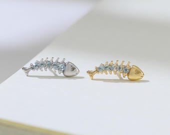 May include: Two fish bone-shaped earrings. One earring is silver-toned with light blue accents, and the other is gold-toned with similar light blue accents. The earrings are placed on a white surface.