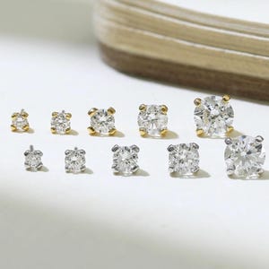 May include: A collection of ten round-cut stud earrings arranged in two rows. The top row features five gold-tone settings with clear stones, while the bottom row features five silver-tone settings with clear stones, all in varying sizes.
